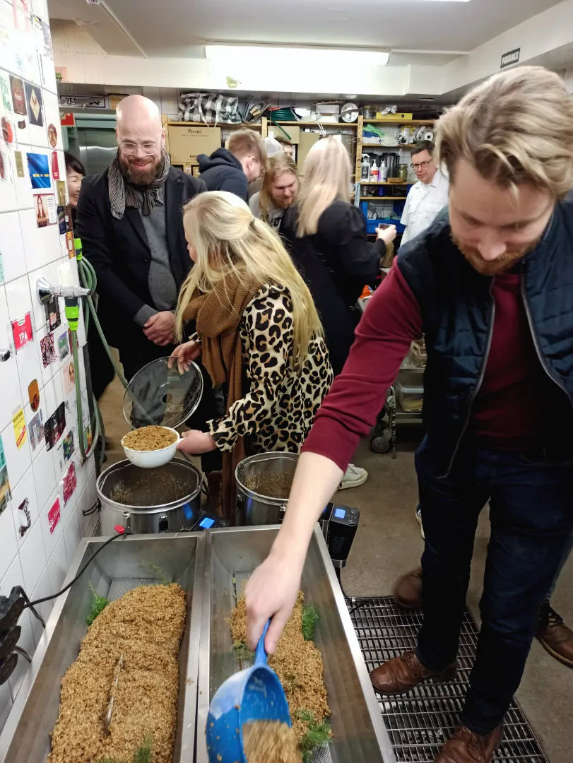 Ryhmän ihmisiä valmistamassa olutta kellaritilassa, jossa mies kauhalla lisää mallasta käymisastioihin.