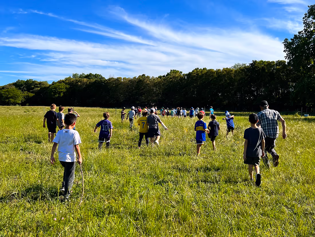 Group of children and an adult walking through a green grassy field toward a tree line under a blue sky with white clouds.