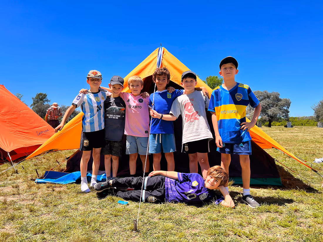 Group of seven boys posing in front of orange tents on grassy field under clear blue sky, one boy lying down in front.