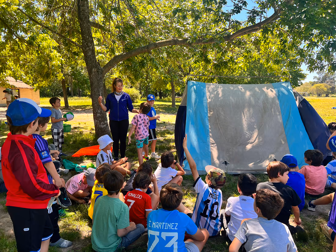 Group of children sitting and standing around a blue and grey tent in a sunny park with trees, while a woman stands nearby speaking.
