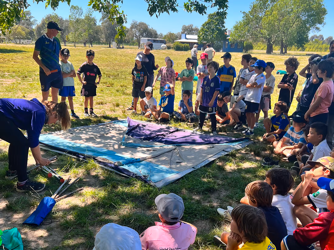 Group of children gathered outdoors in a grassy park listening to adults demonstrating how to assemble a tent.