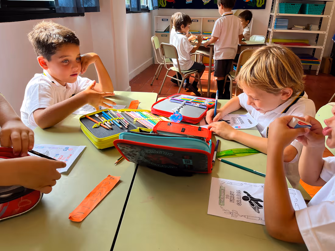Young children in white shirts working on coloring and writing activities at classroom tables with pencil cases and supplies.
