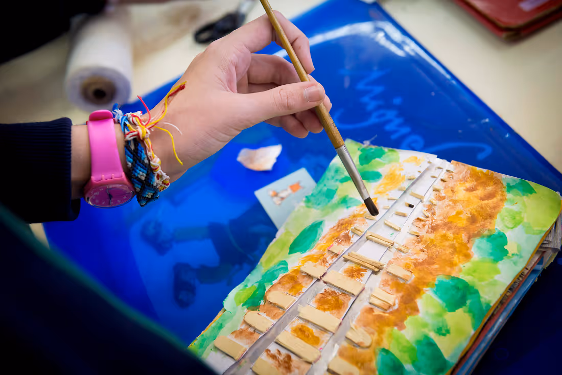 Person painting a detailed, colorful model of a bridge with green and brown shades on paper.