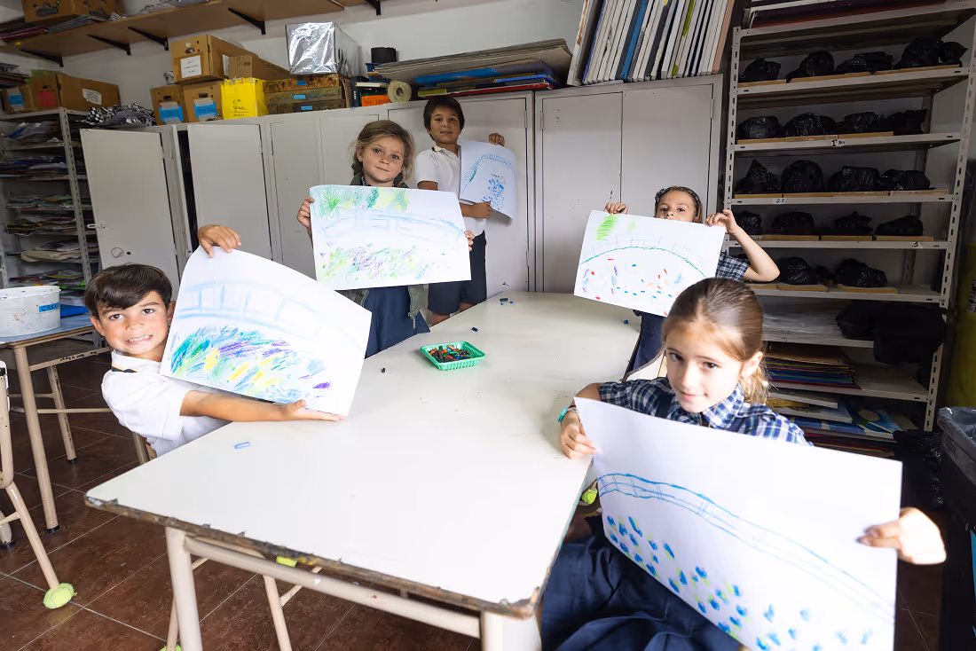 Five children sitting around a classroom table showing colorful drawings on large sheets of paper.