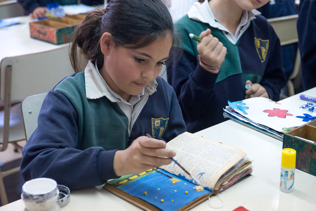 A girl in a school uniform painting a picture in a notebook at a classroom desk.