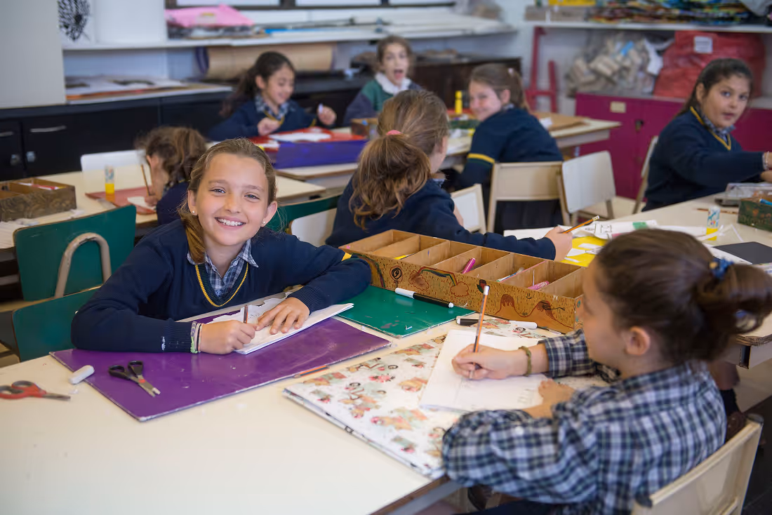 Smiling girl in school uniform sitting at a desk drawing with pencil among classmates in a classroom.