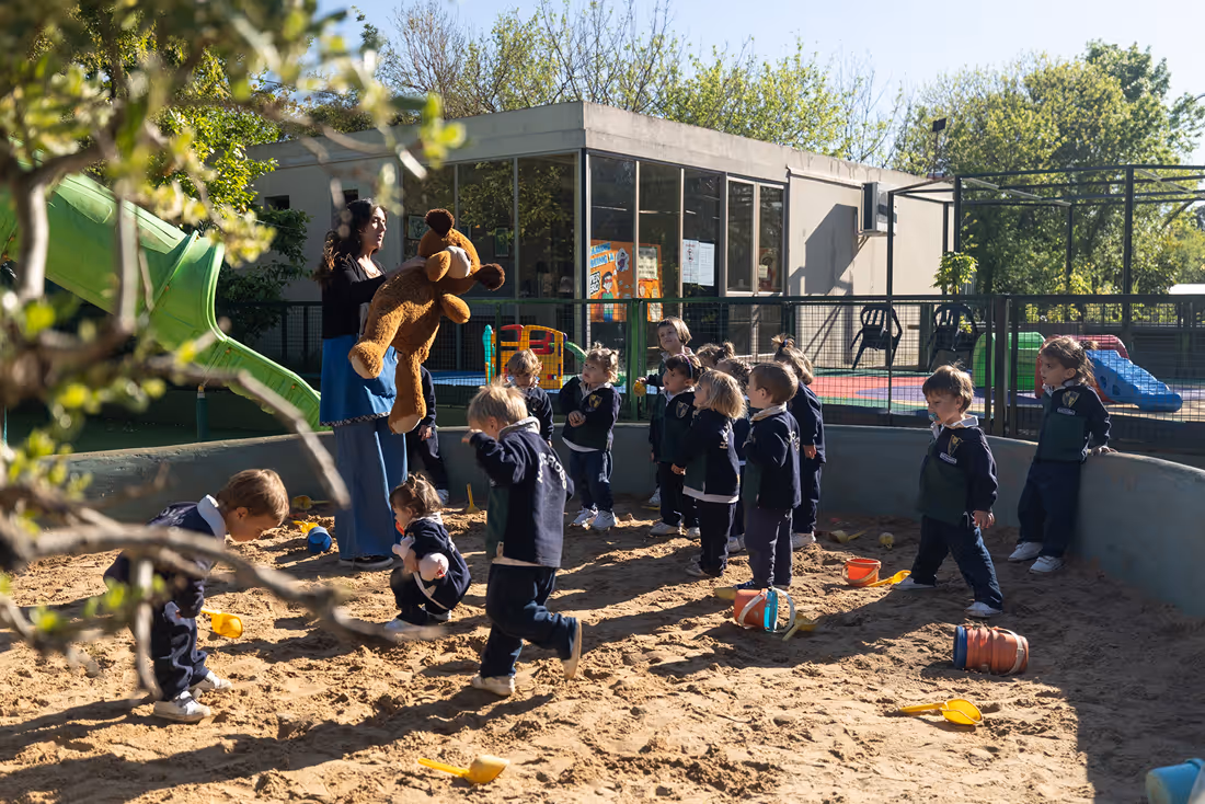 Group of young children playing in a sandpit while a woman holds a large teddy bear near a playground slide.