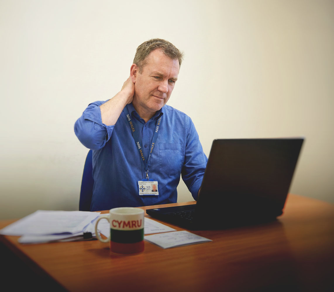 Man in blue shirt with NHS Wales lanyard sitting at a desk using a laptop, with a Cymru mug and papers on the desk.