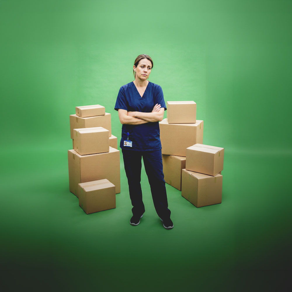Tired healthcare worker in navy scrubs standing with arms crossed surrounded by stacked cardboard boxes on a green background.