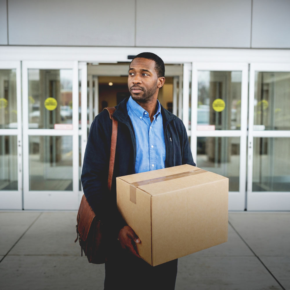Man wearing a blue shirt and black jacket carrying a large cardboard box in front of glass automatic doors.