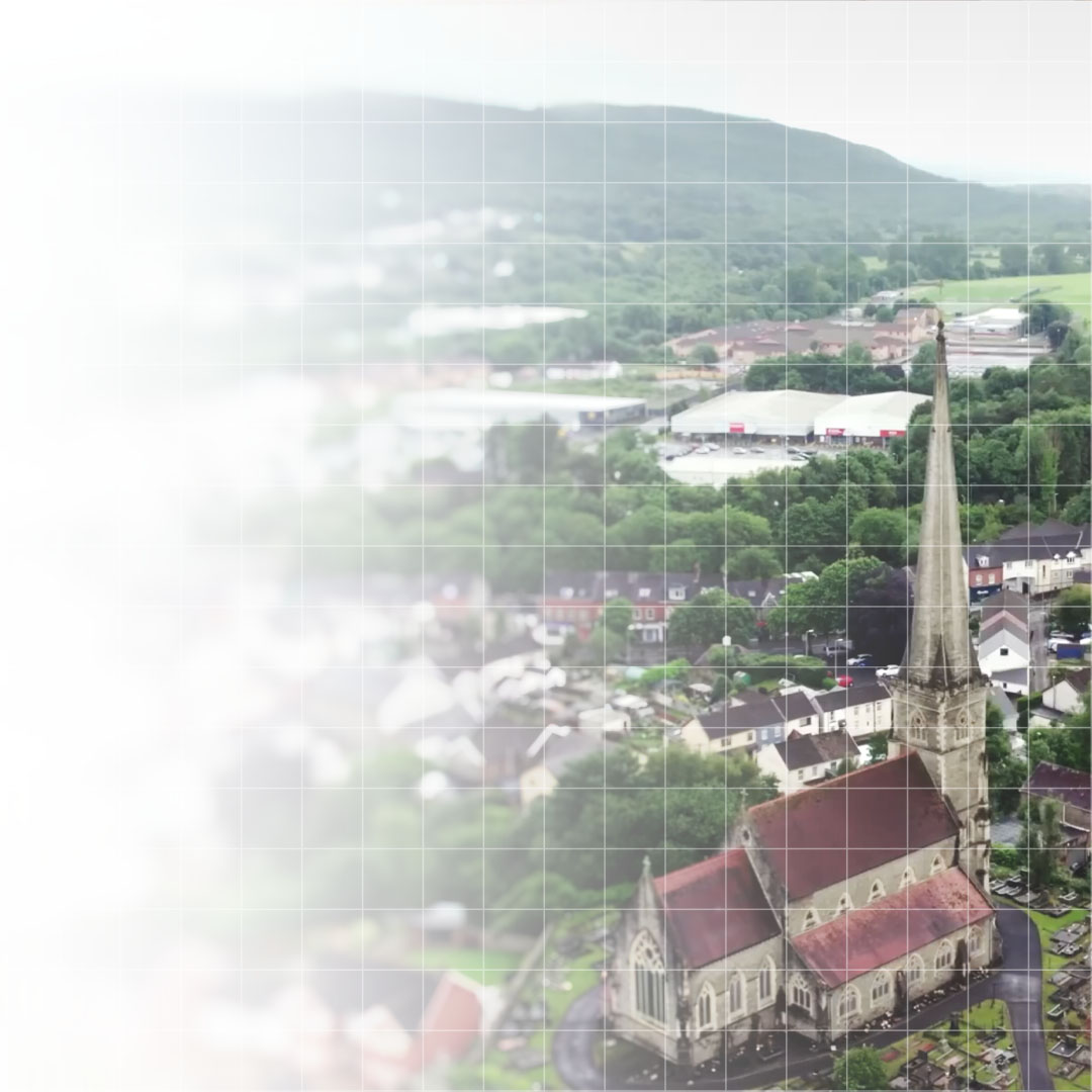 Aerial view of a stone church with a tall spire surrounded by houses, trees, and hills in the background.