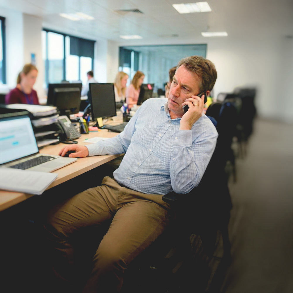 Man in a light blue shirt speaking on a phone in a busy office with coworkers at desks.