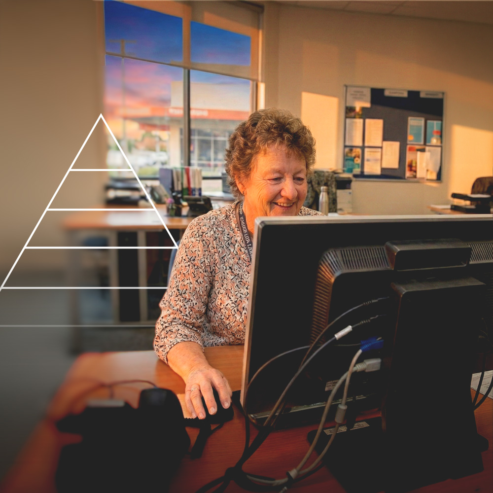 Smiling elderly woman working on a desktop computer in an office with sunset light through the window.