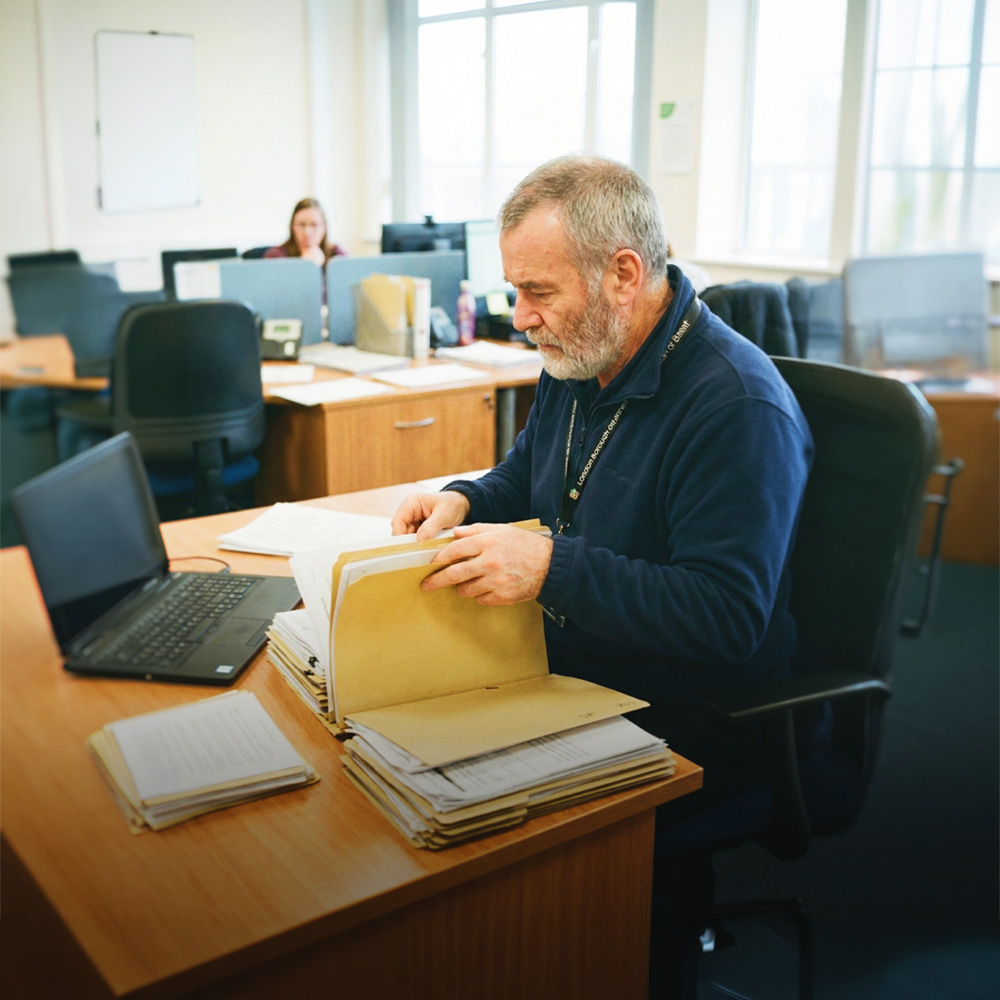An older man with gray hair and beard sorting through file folders at a desk in a bright office.
