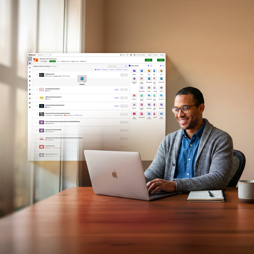 Smiling man wearing glasses and a gray cardigan working on a laptop at a wooden desk with a notebook and mug beside him.