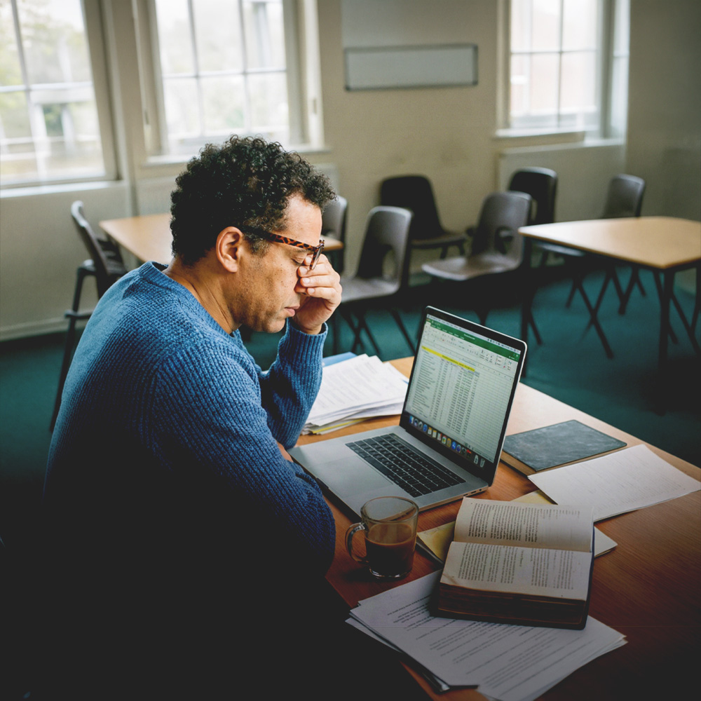 Man in blue sweater sitting at a desk with a laptop displaying a spreadsheet, surrounded by papers and a book, holding his face in his hand, appearing stressed.
