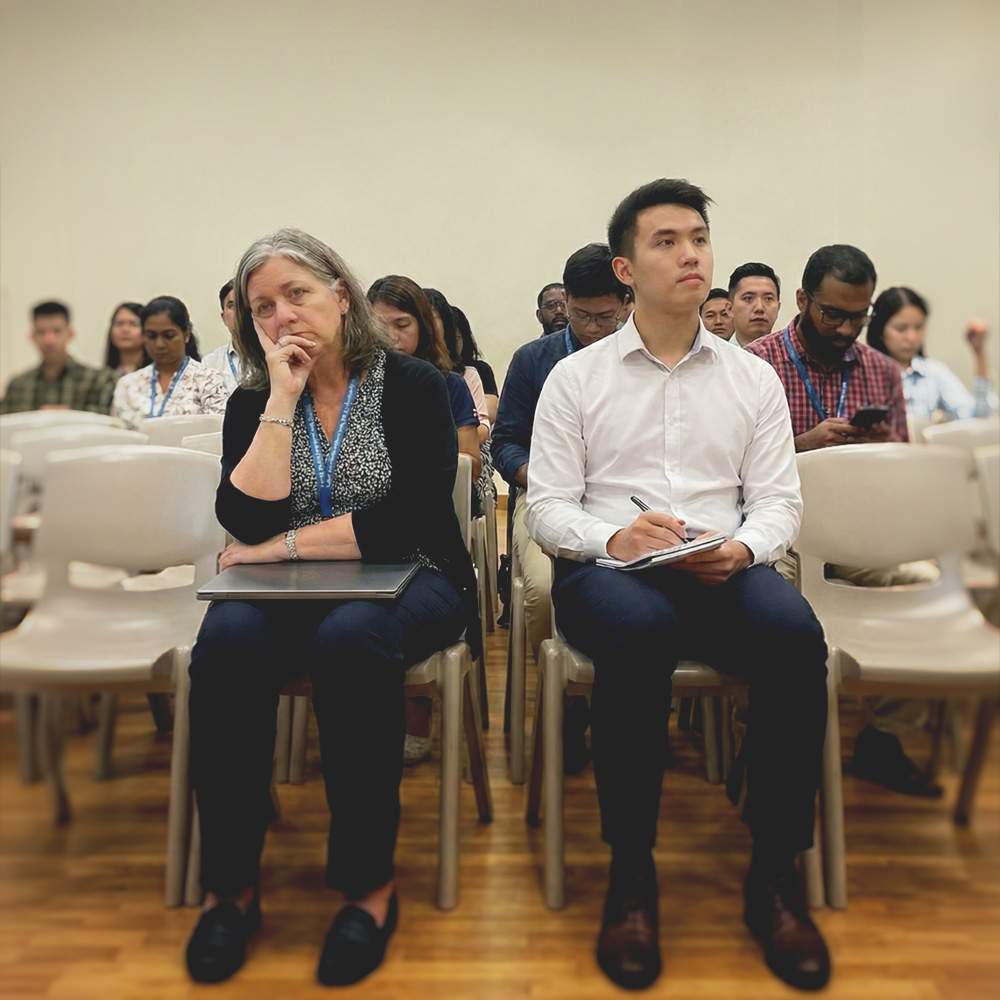 Diverse group of adults seated in a classroom setting, with one woman looking thoughtful and a man taking notes.