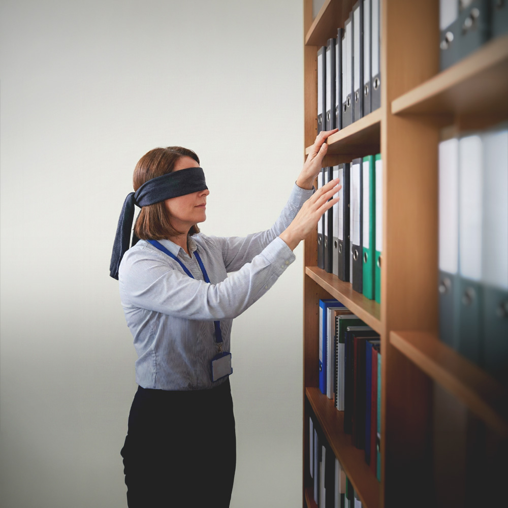 A blindfolded woman wearing a business badge reaches for files on a wooden office shelf.