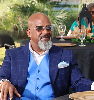 Man with gray beard wearing sunglasses, a blue three-piece suit, and a white shirt sitting at an outdoor table.