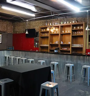 Indoor bar area with metal stools, a corrugated metal counter, exposed brick walls, and wooden shelves holding glassware and bottles.