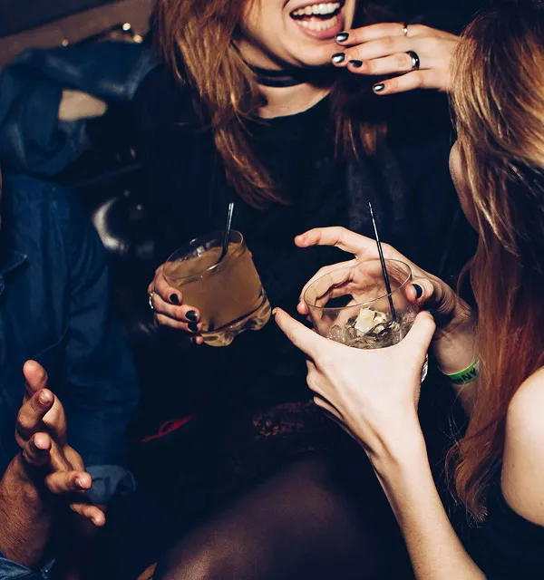 Two women holding drinks and laughing while socializing in a dimly lit setting.