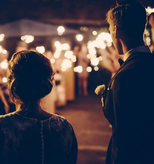 Bride and groom seen from behind walking through a dimly lit path with guests holding sparklers.