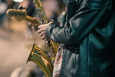 Close-up of a musician in a leather jacket playing a golden saxophone with another musician holding a guitar in the blurred background.