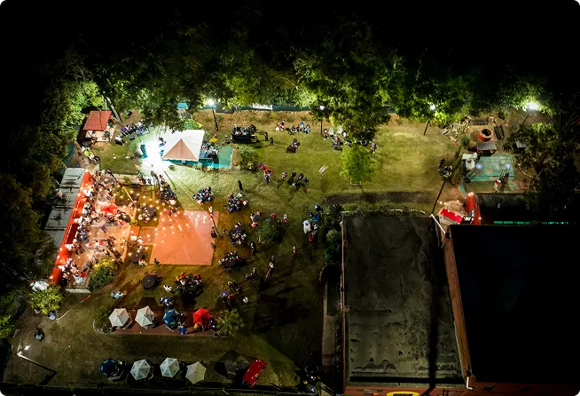 Aerial nighttime view of an outdoor event with groups of people seated on chairs and blankets under string lights near tents and booths.