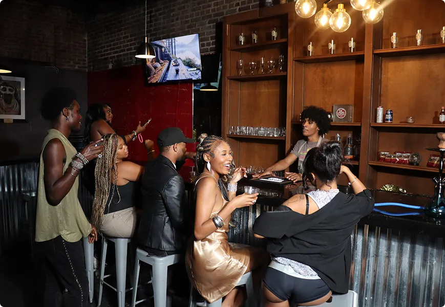 Group of six people socializing at a bar with drinks; bartender serves behind the wooden shelves with glassware.