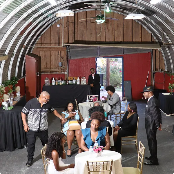 Group of people dressed formally sitting and standing around tables with floral centerpieces inside a large, curved metal and wood structure.