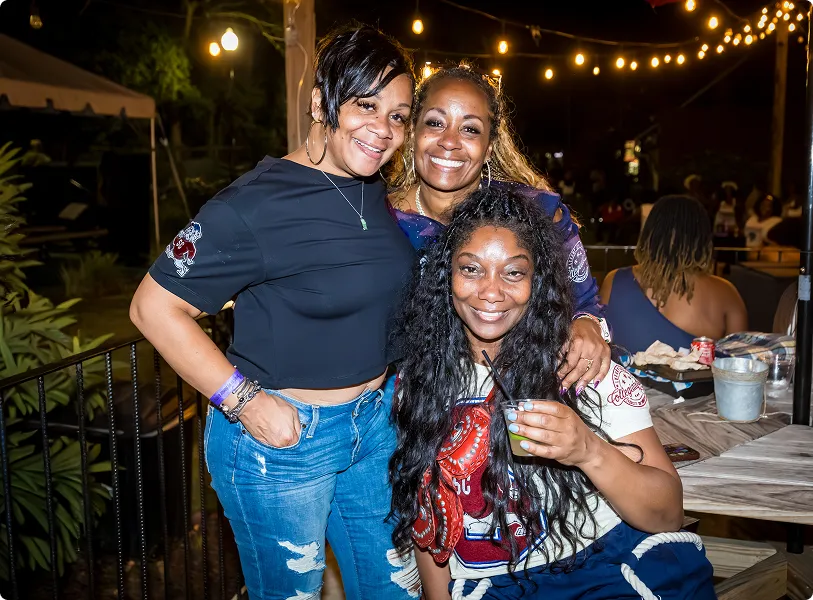 Three women smiling together outdoors at night with string lights in the background, one holding a drink.