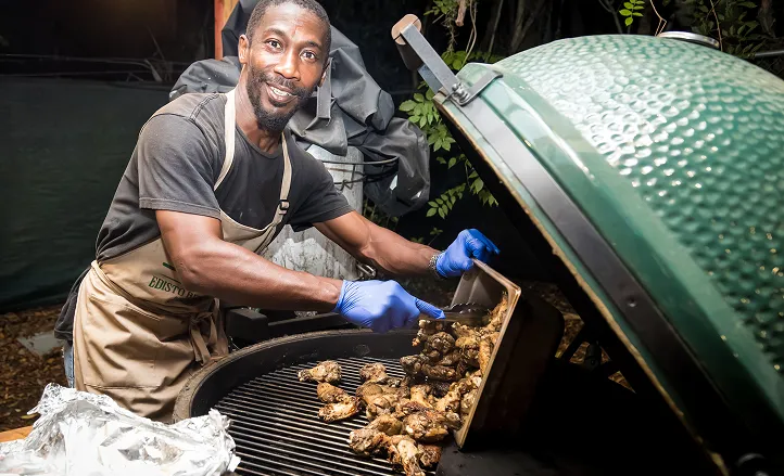 Smiling man wearing apron and gloves grilling chicken wings on a large green barbecue grill outdoors at night.