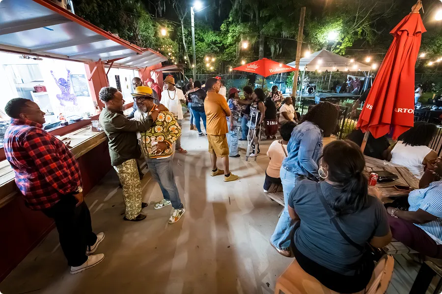 Diverse group of people socializing outdoors at night under string lights near red umbrellas and a food stand.