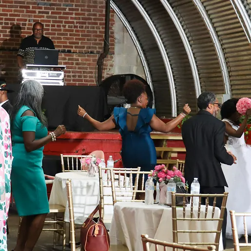 People dancing near tables decorated with pink flowers and water bottles at an indoor event with a DJ playing music.
