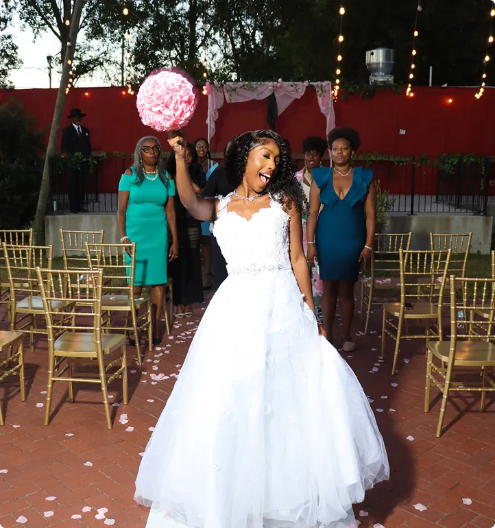 Bride in a white wedding dress holding a pink bouquet joyfully with bridesmaids standing behind her outdoors.