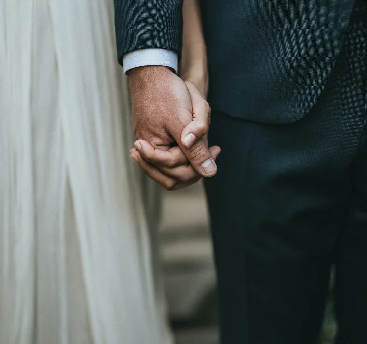 Close-up of a couple holding hands, with the person on the left wearing a white dress and the person on the right wearing a dark suit.
