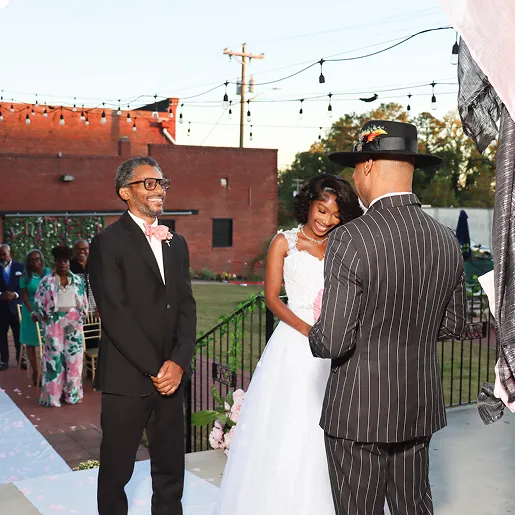 Bride in white wedding dress smiling, standing between two men in suits during an outdoor ceremony.