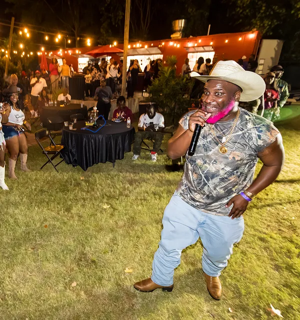 Man with a white cowboy hat and pink beard singing outdoors at night with a crowd and string lights in the background.