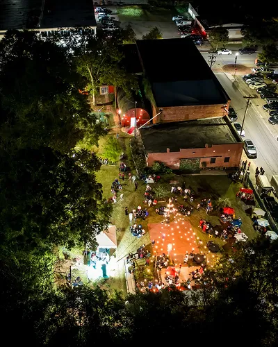 Aerial night view of a crowd gathered outdoors near a building lit by string lights and spotlights in a park or event space.