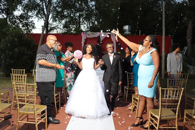 Newlywed couple walking hand in hand down the aisle outdoors, surrounded by guests throwing flower petals.