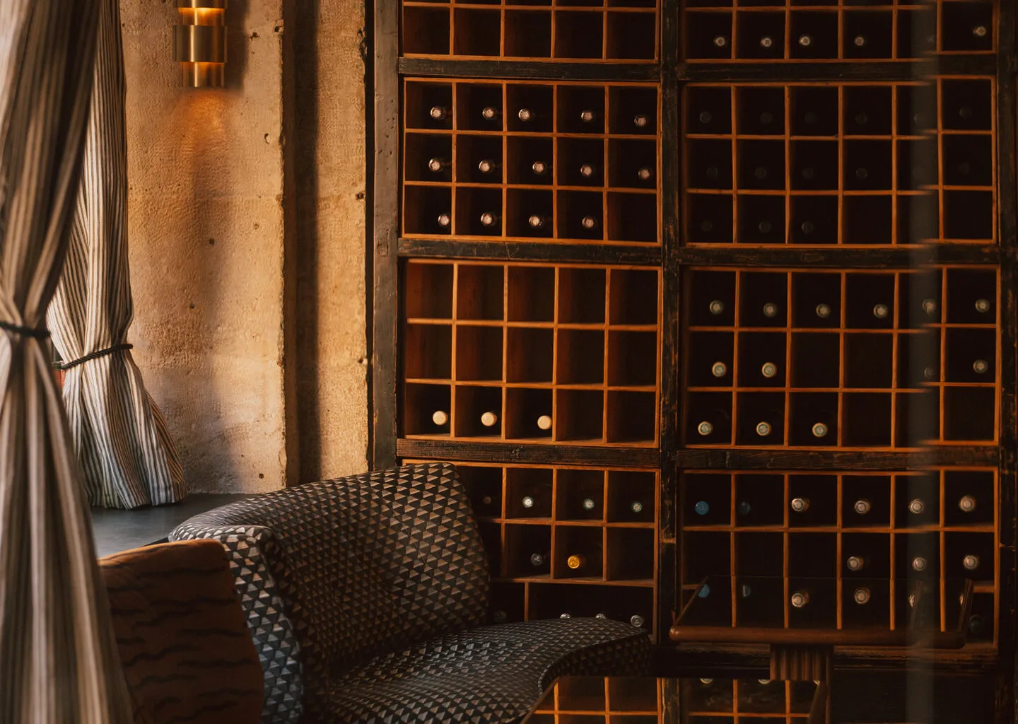 Cozy corner with patterned sofa, wooden wine rack filled with bottles, striped curtains, and a small table.