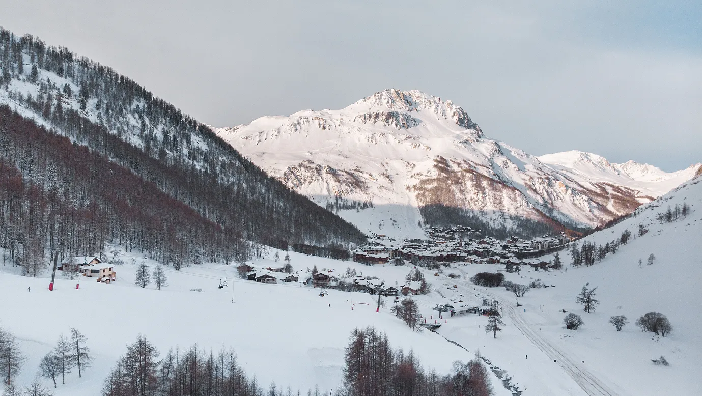 Snow-covered mountain village with ski lifts and leafless trees under a partly cloudy sky.