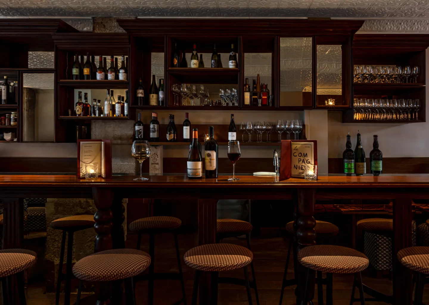 Cozy wine bar interior with wooden counter, wine bottles, glasses, menus, and cushioned stools under warm lighting.