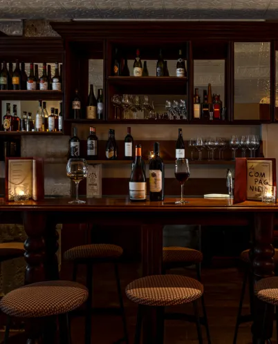 Dimly lit bar with wooden stools, bottles of wine and liquor on shelves, and glasses on the counter.
