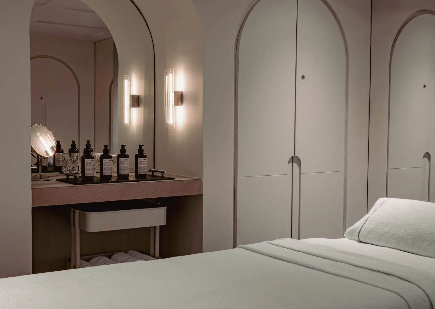 Minimalist spa room with a neatly made treatment bed, a vanity with skincare bottles, a round mirror, and soft wall lighting.