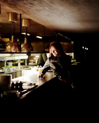 Woman sitting alone at a dimly lit bar counter holding a cocktail glass and smiling.