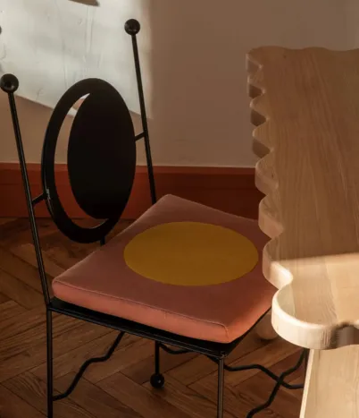 Chaise en métal avec dossier circulaire noir et coussin d'assise rose orné d'un grand cercle jaune, à côté d'une table en bois au bord festonné.