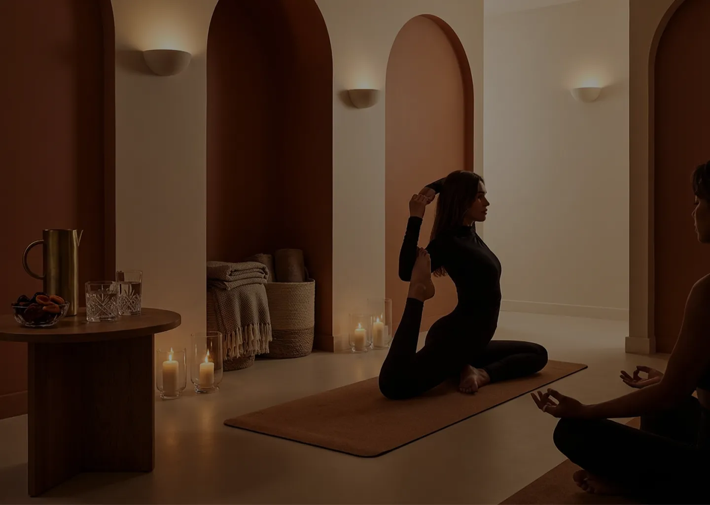Two women practicing yoga in a softly lit room with arched wall niches, candles, and cozy decor, one performing a seated backbend pose and the other meditating.