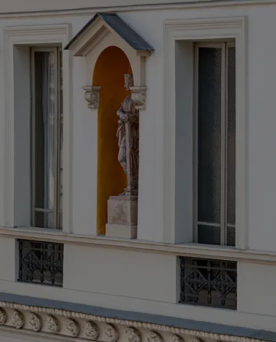 Stone statue of a figure in a niche between two windows on a light-colored building facade.