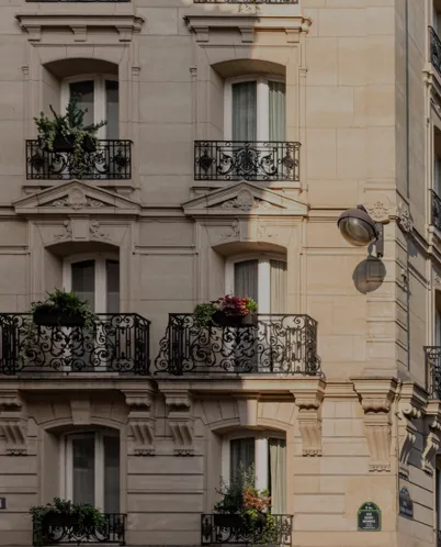 Beige stone Parisian building facade with ornate iron balconies and plants on each level, lit by warm sunlight.